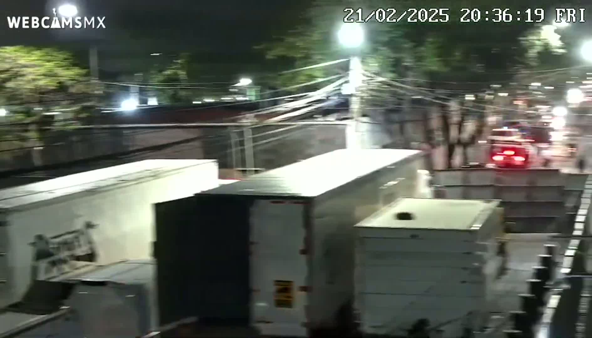 At night, several large, light-colored trucks are parked or stopped near a chain-link fence, with a street and moving vehicles visible in the background.