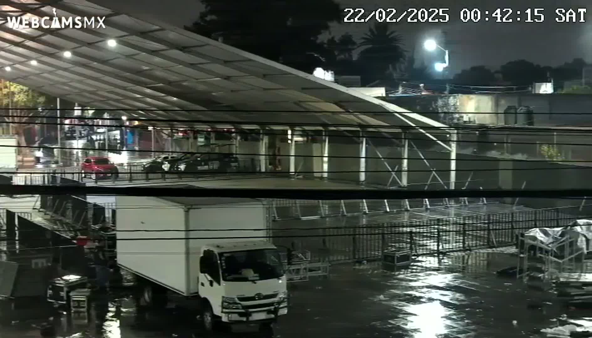 A white box truck is parked in a rain-soaked lot under a large metal awning at night, with several other vehicles visible in the background.