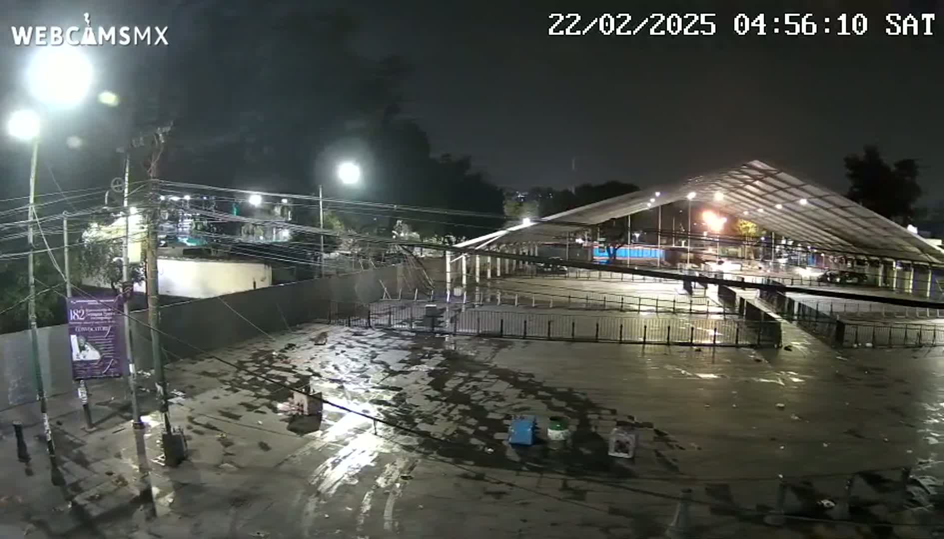 A nighttime, wet outdoor scene shows a large, empty paved area under a metal-framed covered structure, surrounded by fences, trees, and overhead power lines.