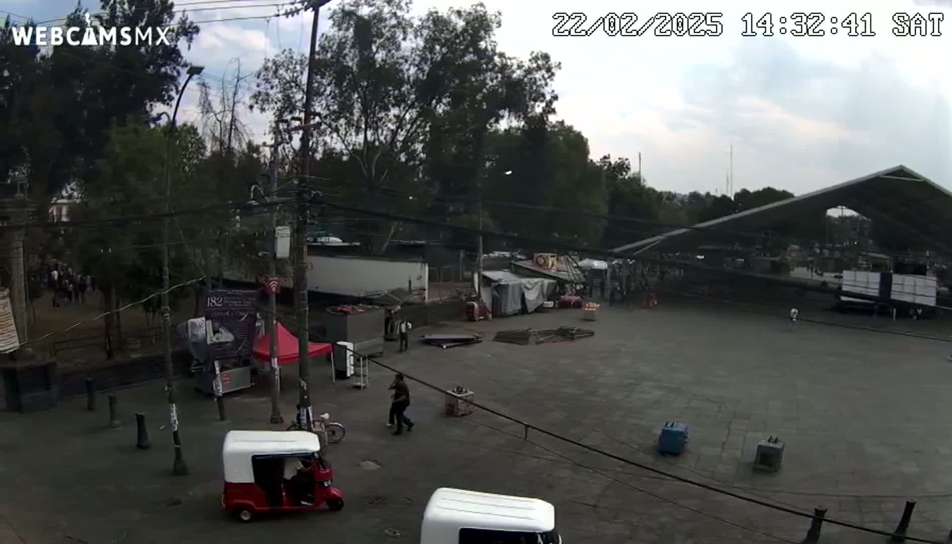A paved plaza under an overcast sky features several small stalls and vendors, a large covered structure, and a few people walking around, with some three-wheeled vehicles parked nearby.