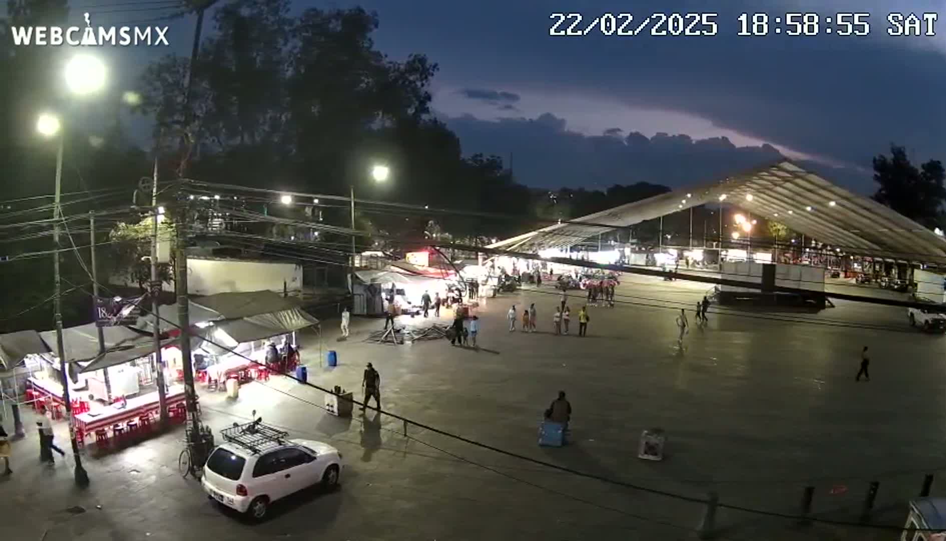 An outdoor nighttime scene shows a large, open-air market with numerous food stalls and people walking around under a partly cloudy sky.