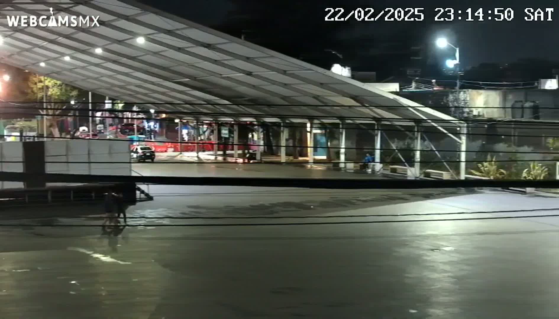 At night, a mostly empty plaza sits under a large, covered structure, with a few people visible.