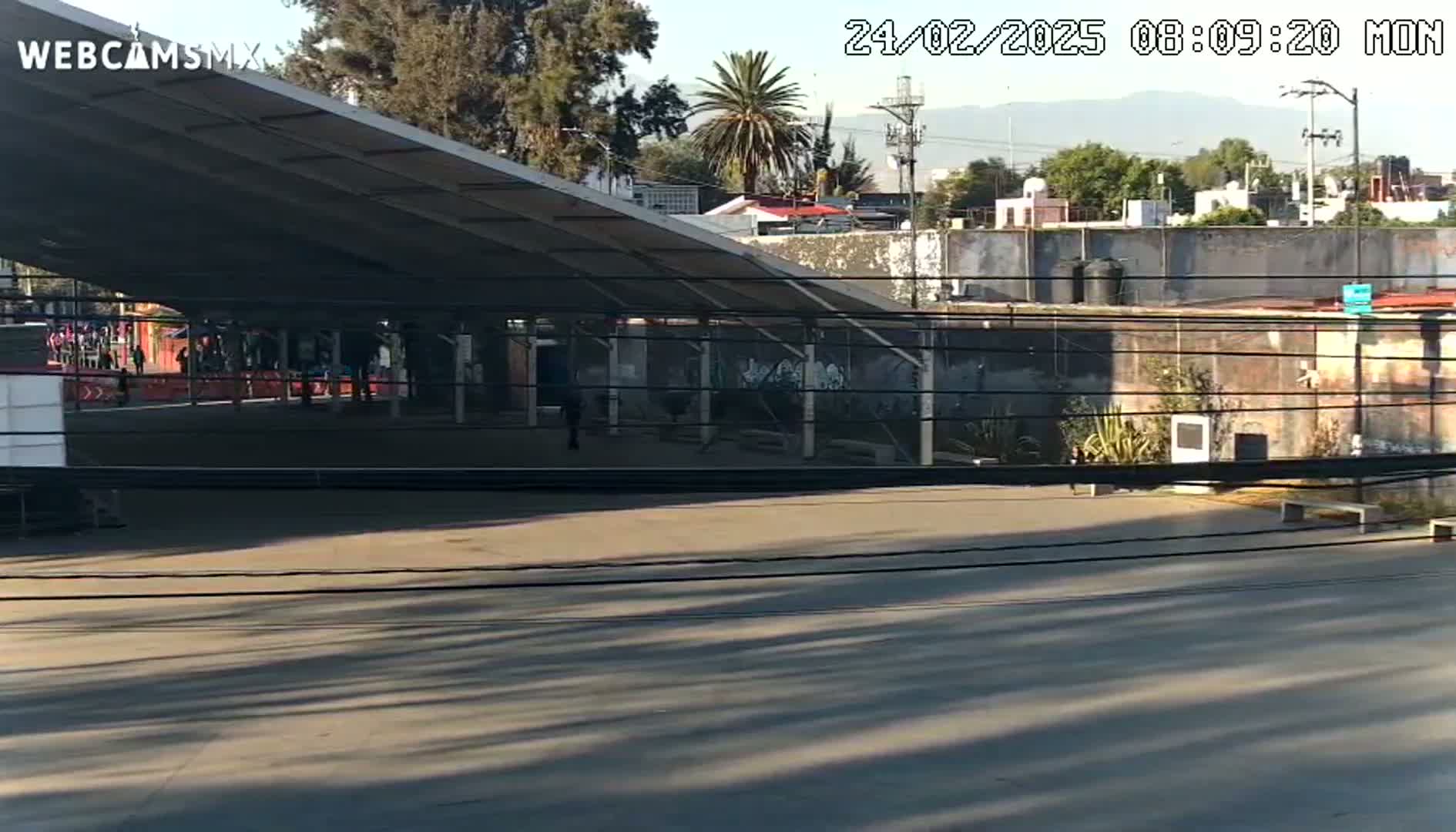 A mostly empty paved area sits under a large metal awning, with a few people visible in the distance, under a sunny sky.
