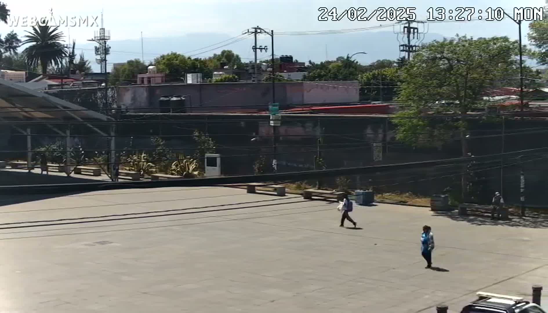 A mostly empty paved plaza is visible, surrounded by buildings under a sunny sky, with a few people walking around.