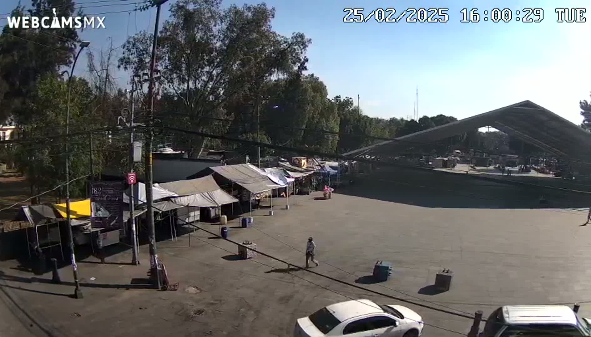 A paved area with several market stalls under tents and a large covered structure, surrounded by trees under a clear, sunny sky.
