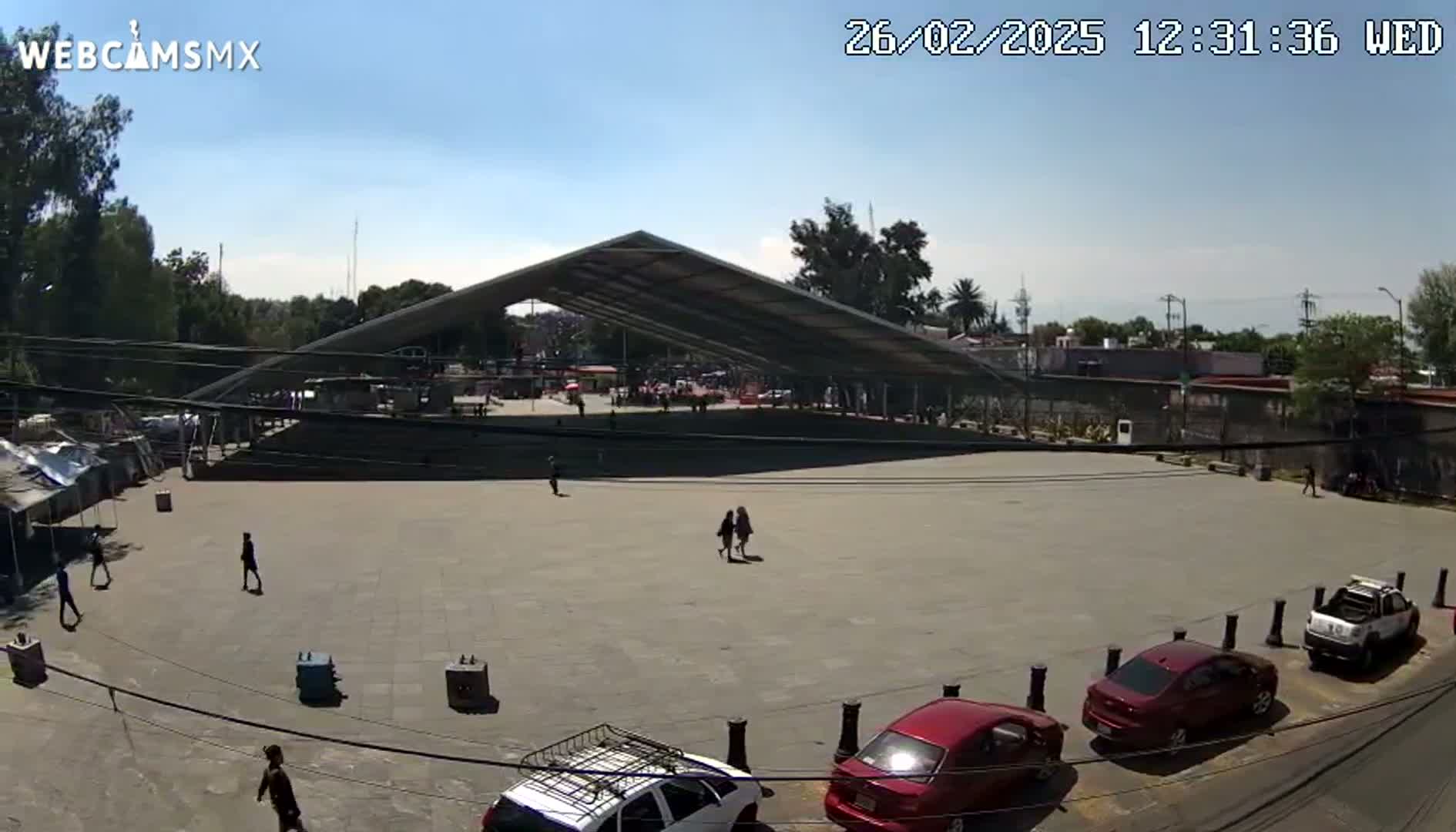 A large, open plaza with a modern, peaked roof structure, several cars parked nearby, and a few people walking around under a sunny sky.