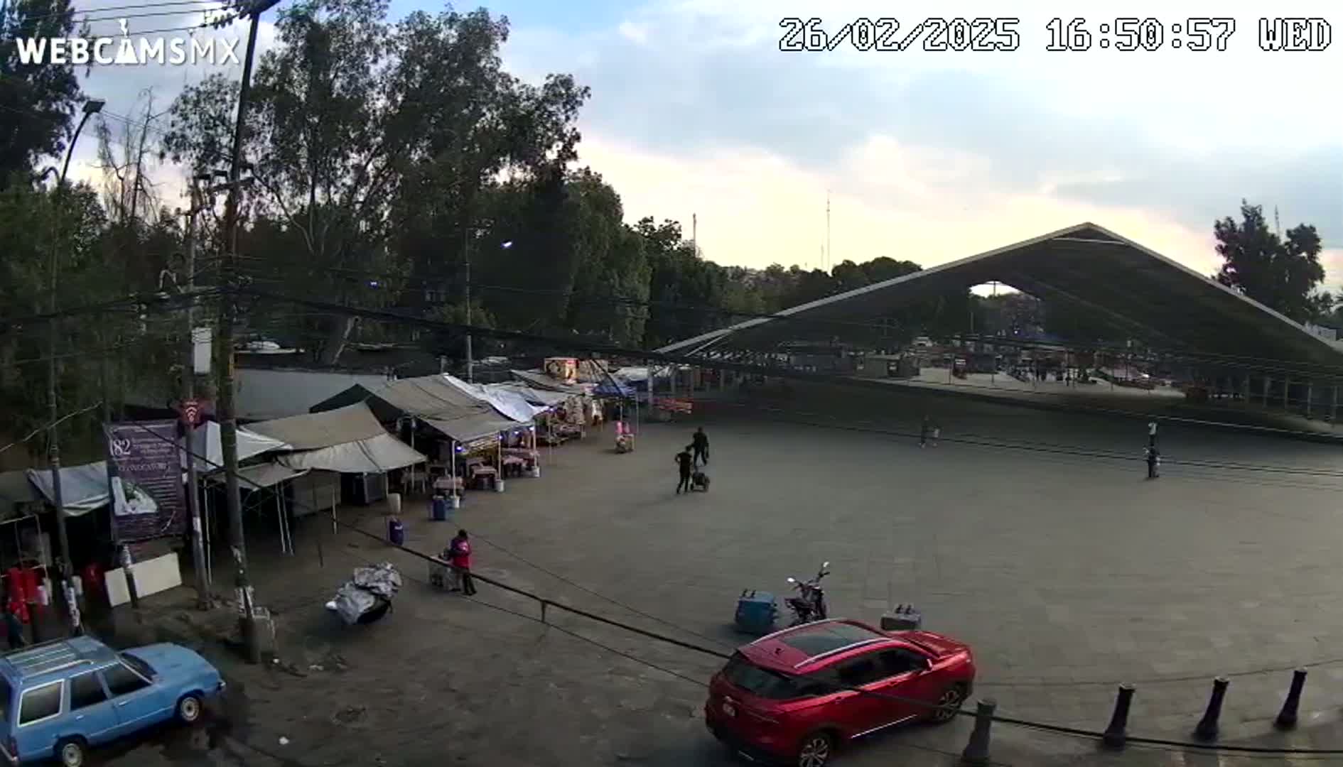 An overcast day reveals an outdoor market area with numerous tents and a large, modern pavilion, several vehicles, and a few people scattered throughout.