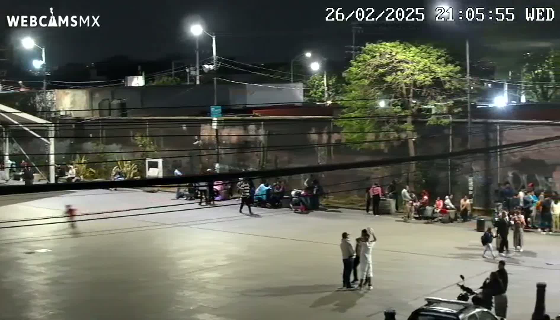 At night, under artificial light, numerous people are gathered in an open-air plaza near buildings, some sitting and others standing, with a few people walking or riding a scooter.