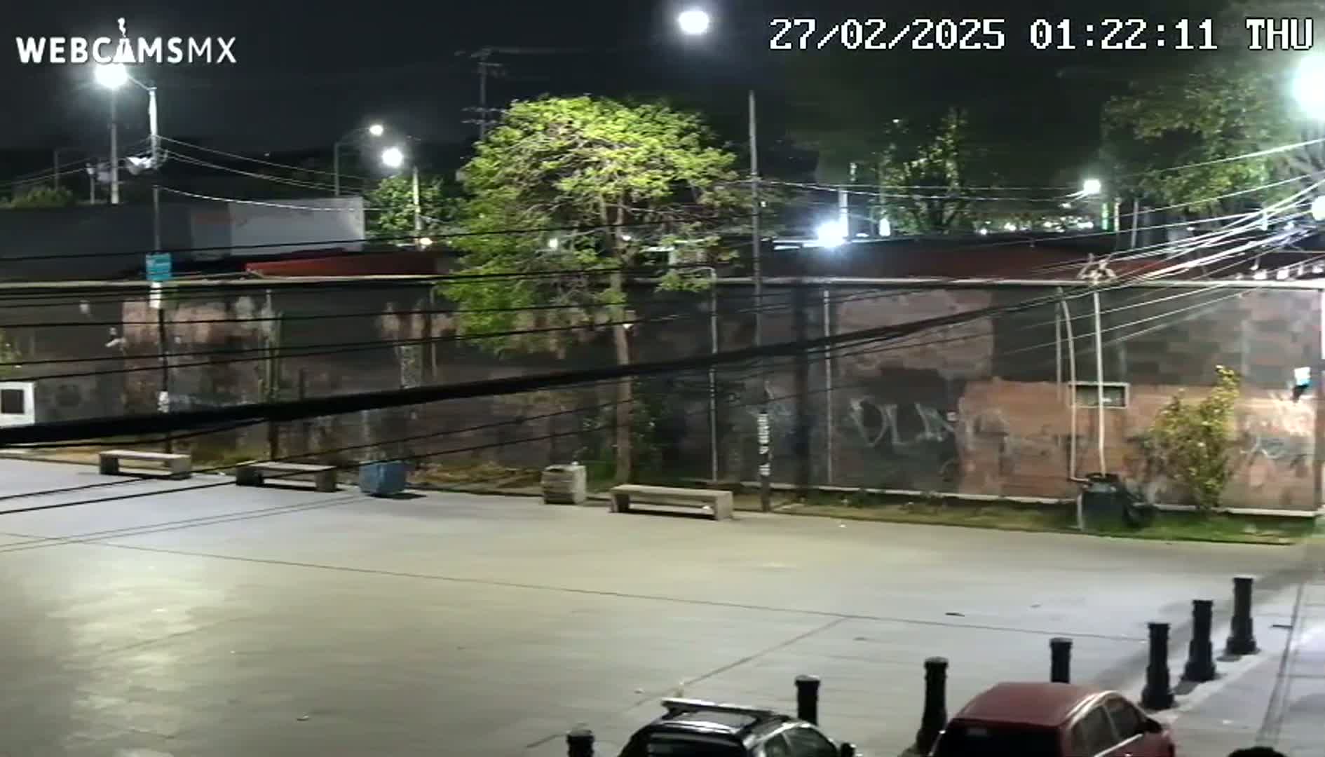 A nighttime, outdoor view of a mostly empty paved area with a few benches, some cars, and a graffiti-covered wall.