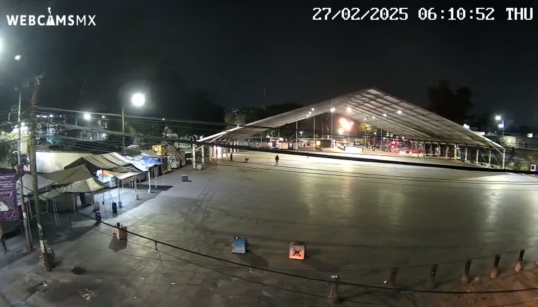 A large, empty plaza at night, illuminated by artificial lights, features a covered structure and several small tents along one side.