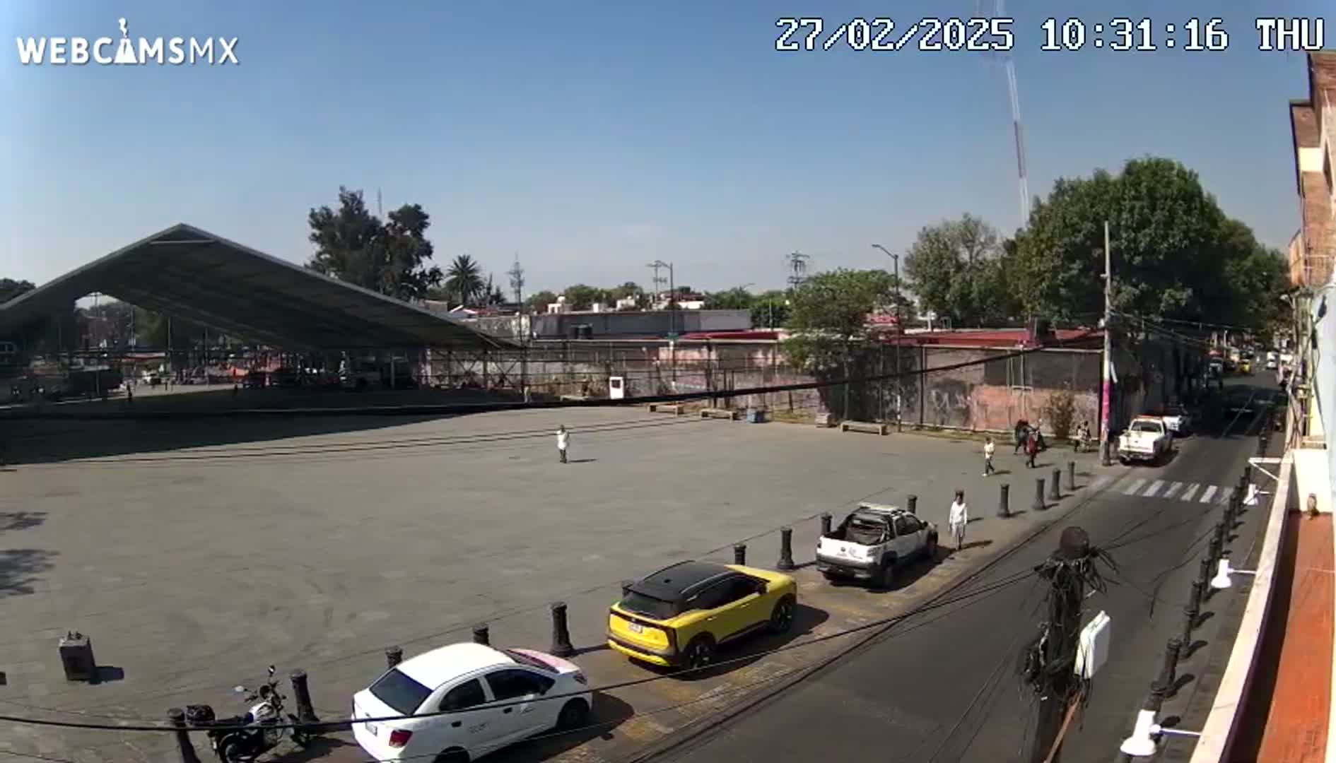 A large, empty plaza under a partly covered structure is seen on a sunny day, with several cars parked nearby and a few pedestrians walking on a street.