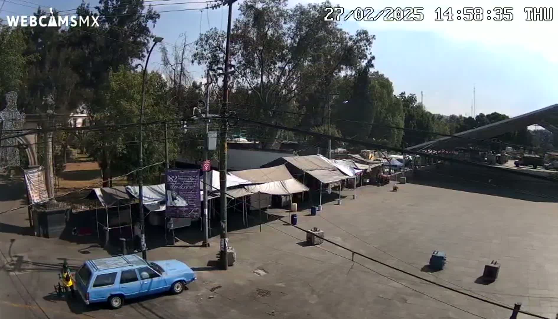 A mostly sunny day reveals a paved area with several temporary market stalls and a light blue station wagon parked nearby, all situated under a canopy of leafy trees.