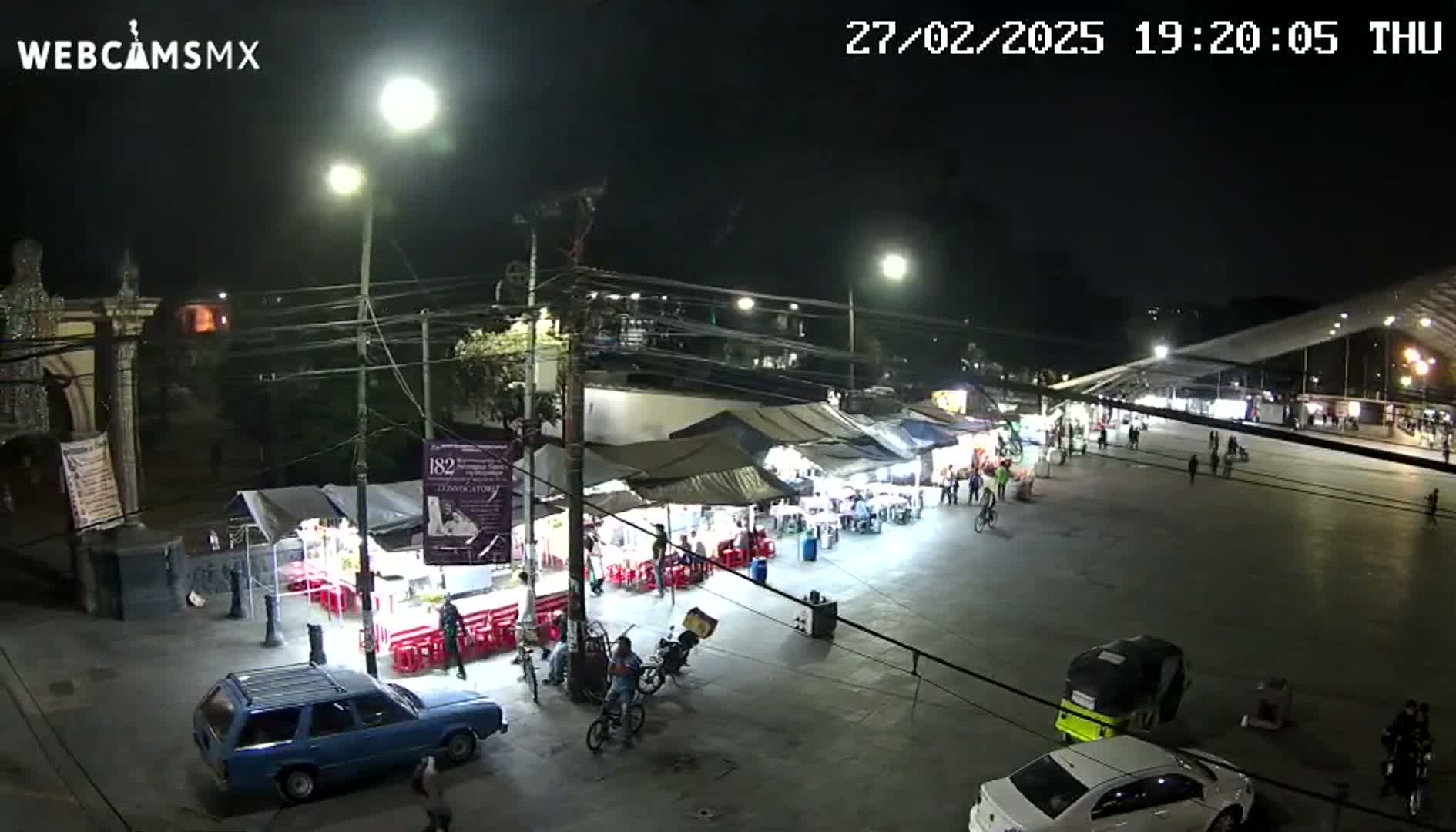 A nighttime, brightly lit outdoor scene shows a plaza with numerous food stalls, people walking, and vehicles parked, under a clear sky.