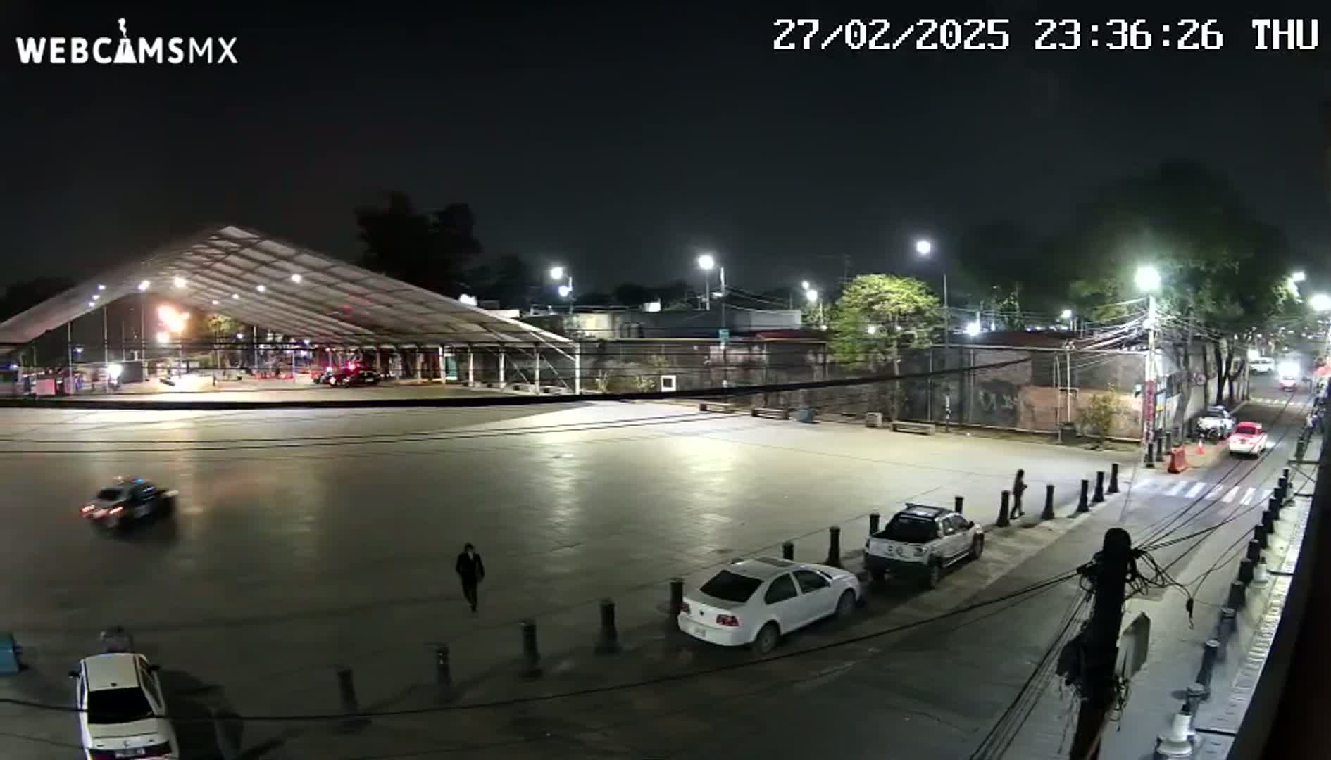 A nighttime, outdoor view of a mostly empty plaza with several cars parked and a large, white tent structure in the background, under well-lit conditions.