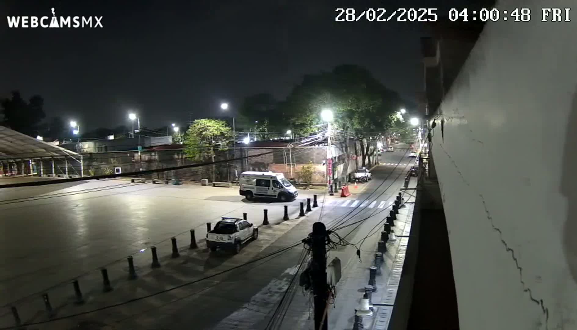 A nighttime, high-angle view of a mostly empty plaza and street, with a few parked vehicles, including a van and a pickup truck, under streetlights.