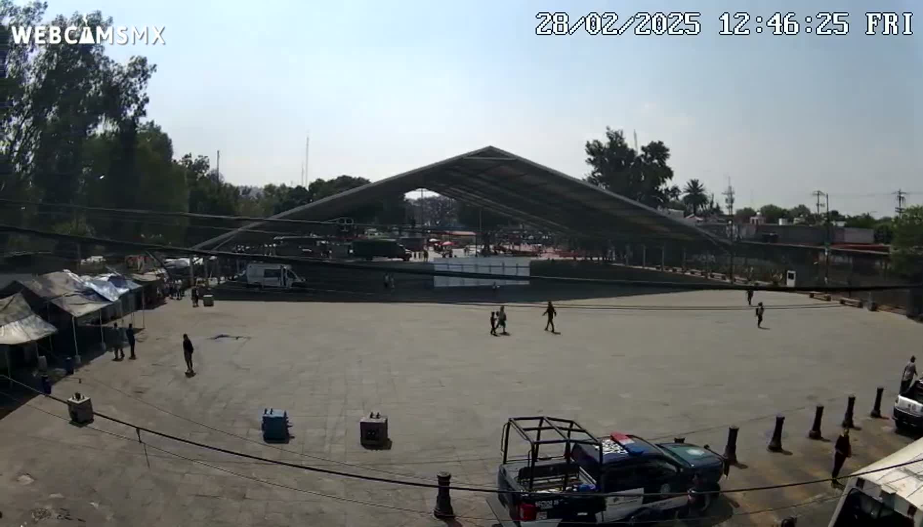 A large, open paved area with a few people and vehicles, including police cars, under a large metal awning on a sunny day.