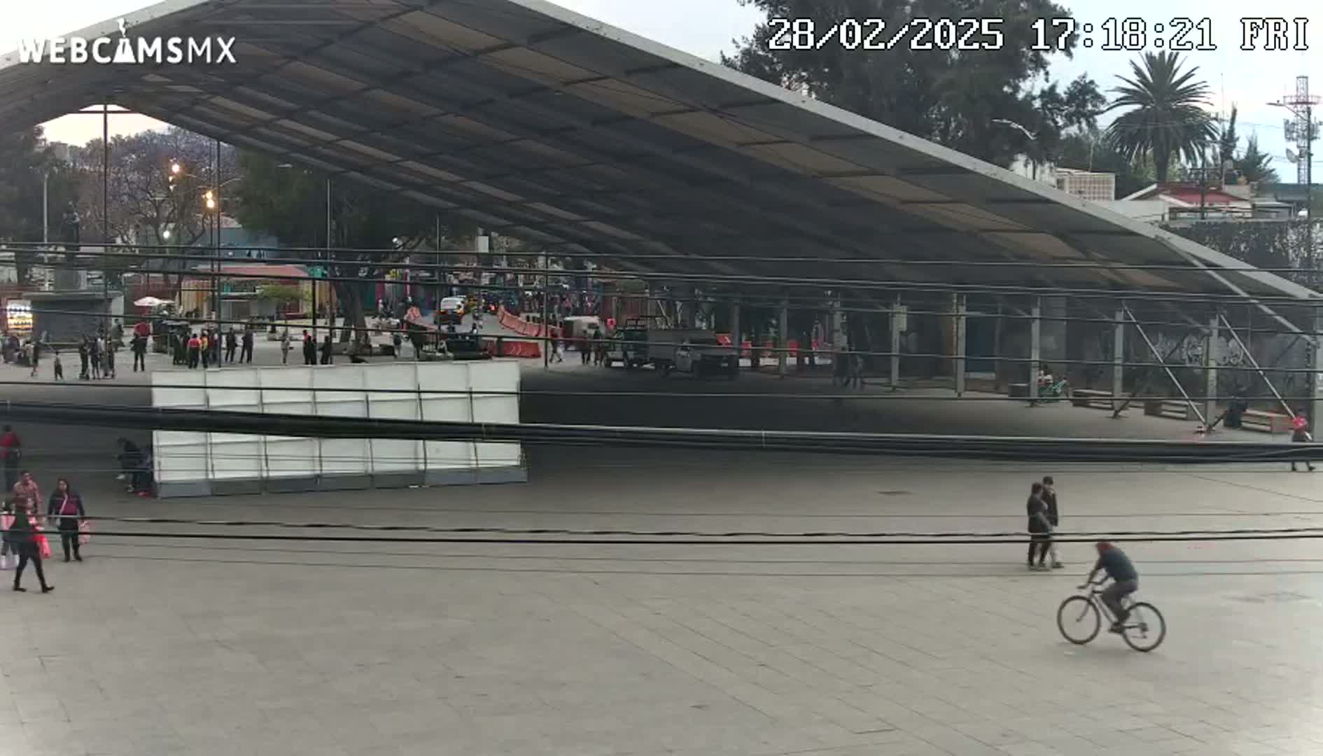 A mostly empty plaza, under an overcast sky, features a large metal structure, several pedestrians, and a person riding a bicycle.