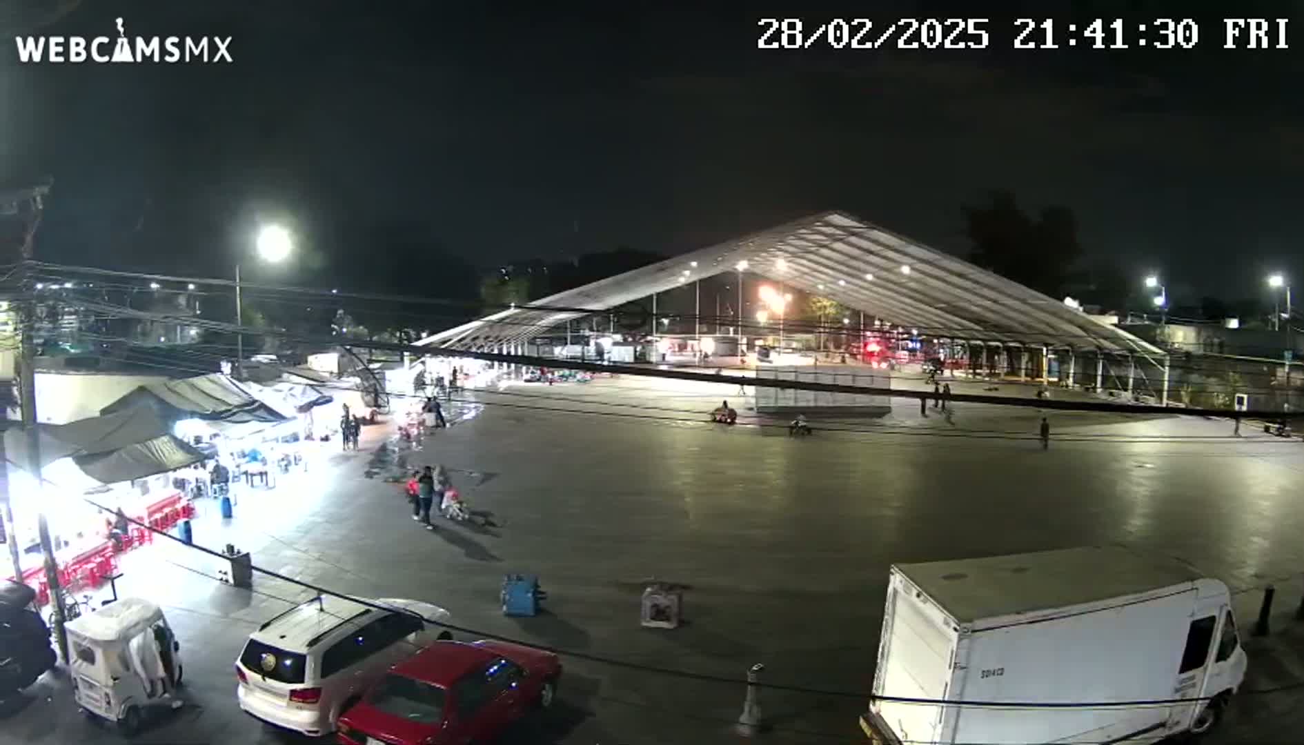 A nighttime, outdoor scene shows a large, open plaza with a large tent structure, some cars, and several people milling about.