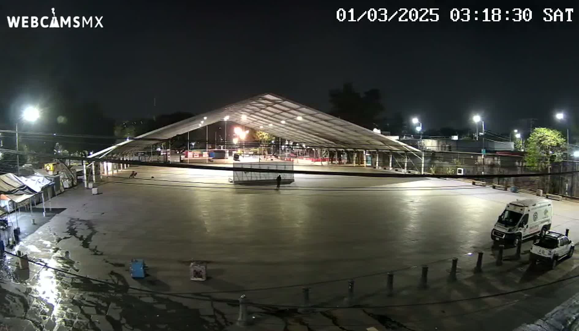 A large, empty, paved area at night under a large, illuminated tent, with an ambulance and a pickup truck parked near the edge, and some small structures and vegetation visible in the background.