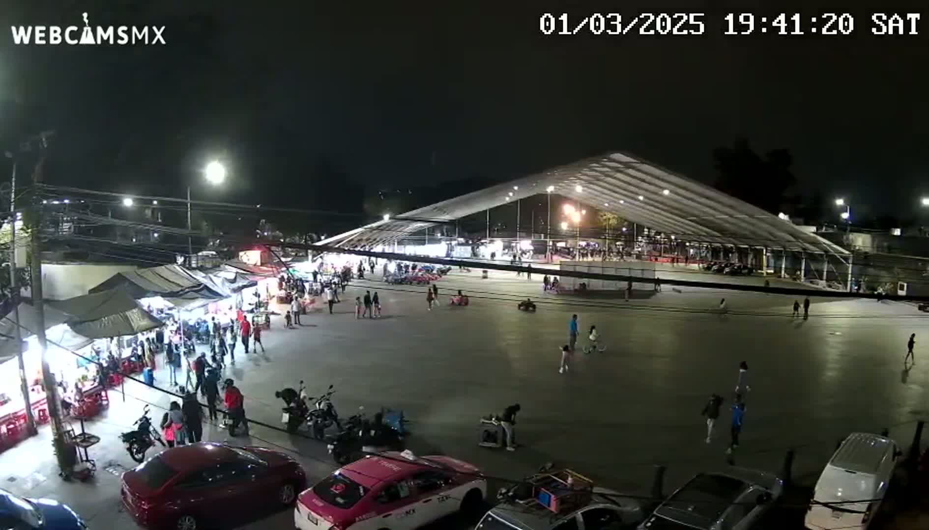 A nighttime outdoor scene shows a large paved area with a few people and parked cars, surrounded by market stalls and a large covered structure.