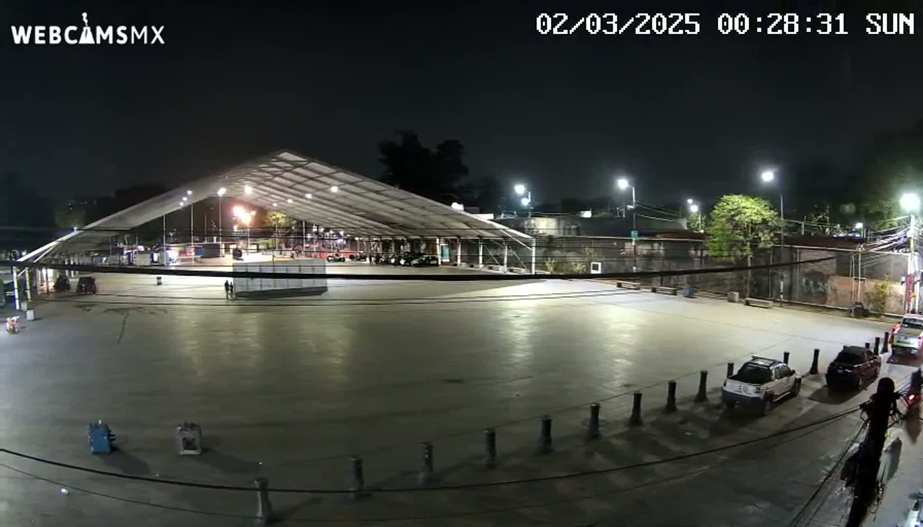 A mostly empty paved plaza at night under a large covered structure, with a few cars parked and some people walking around.