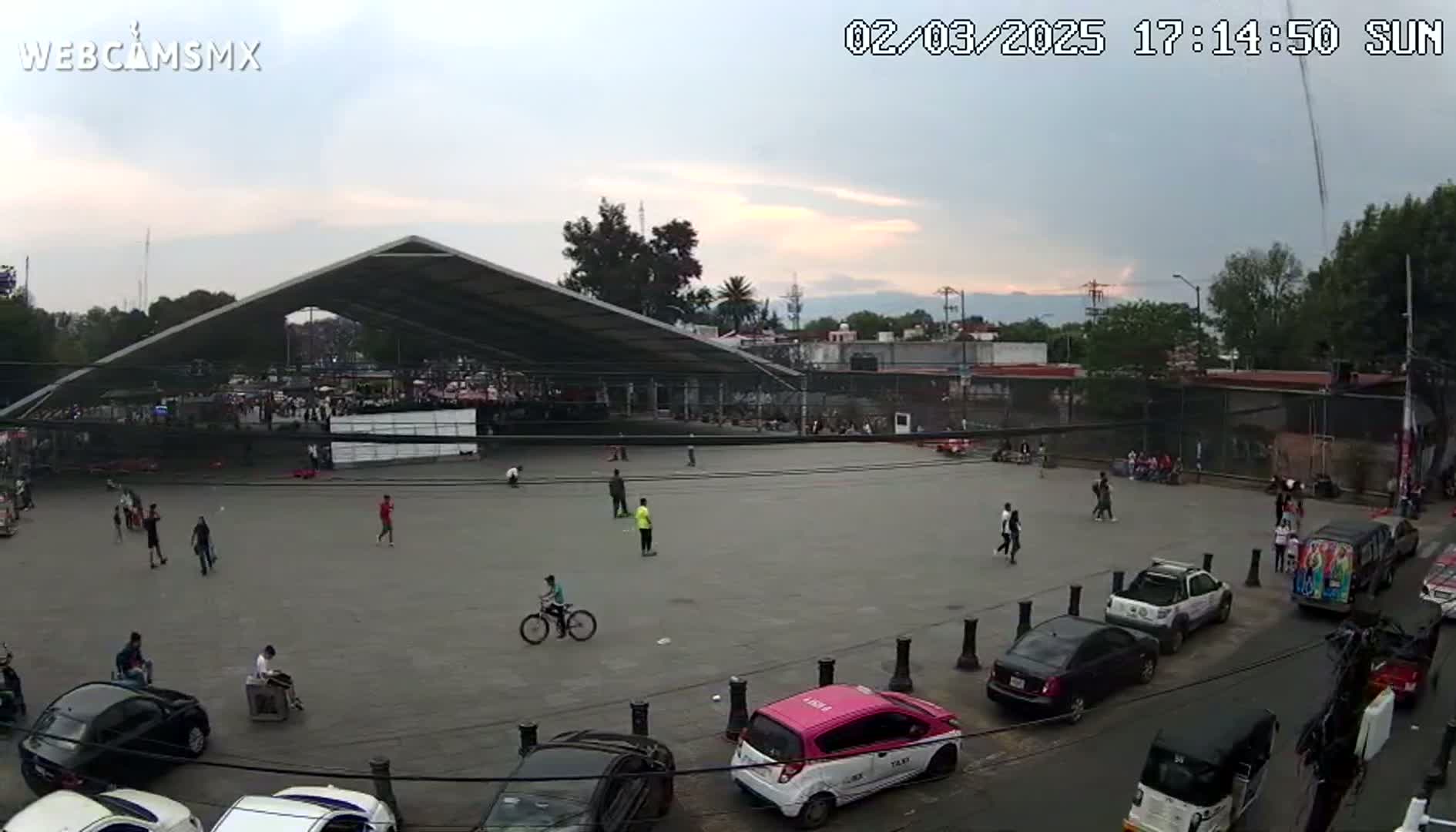 A large paved plaza with a few people playing sports, a large canopy structure, and cars parked along the perimeter under a partly cloudy sunset sky.