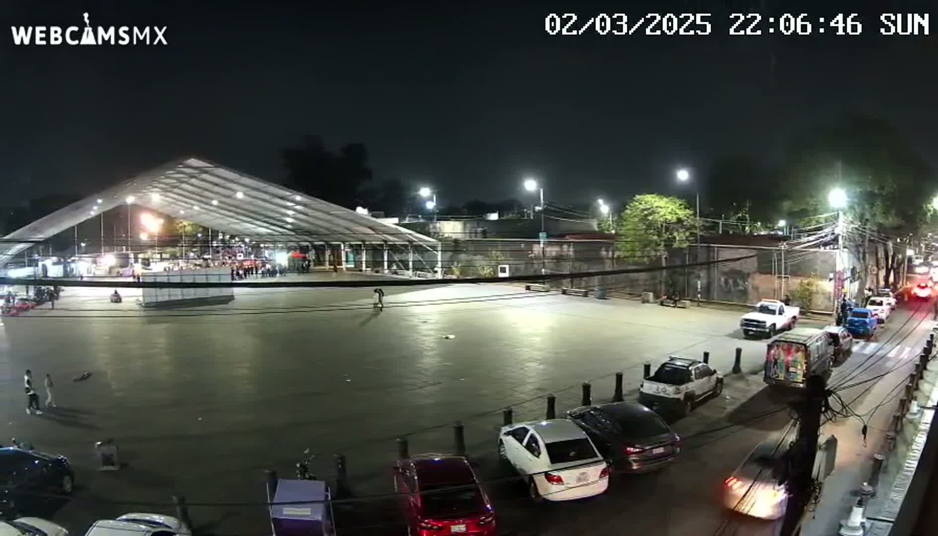 A large, empty plaza at night, illuminated by streetlights, features a large white tent structure, parked cars, and a few pedestrians;  the weather is clear.