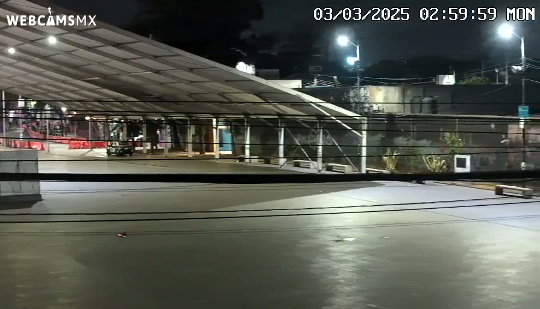 A mostly empty, paved area at night under a large covered structure, illuminated by artificial lights, with a vehicle visible in the distance.