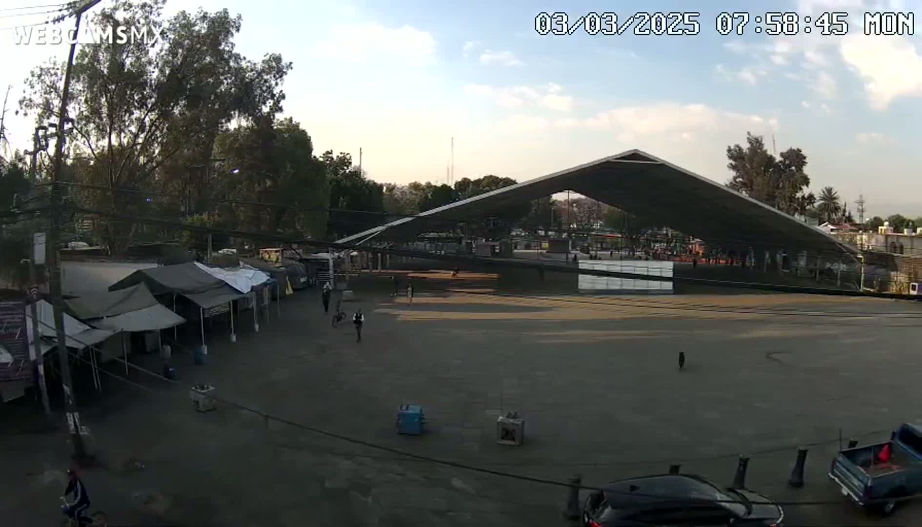 A large, open-air pavilion sits in a mostly empty plaza under a partly cloudy sky, with several small tents and a few people visible near the edges.