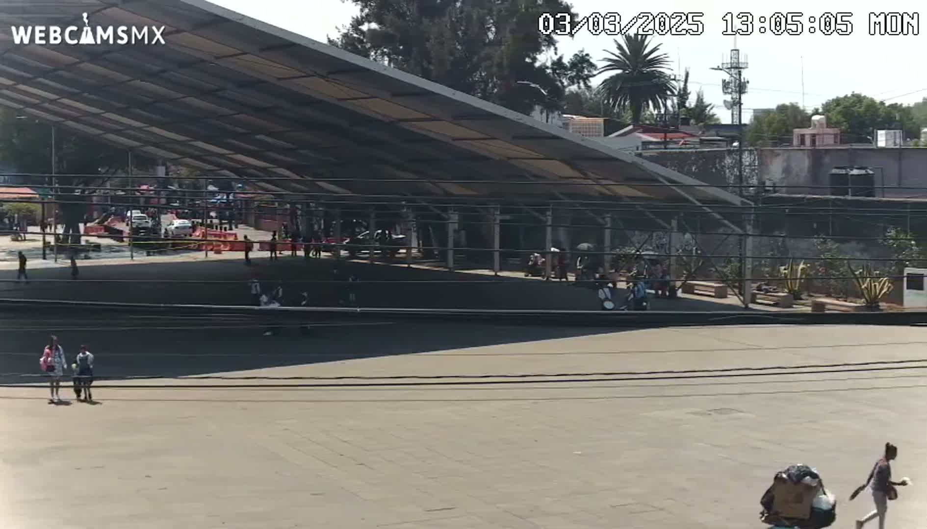 A large, open-air plaza with a modern, angled roof structure is visible under a sunny sky, and several people are scattered throughout the area.
