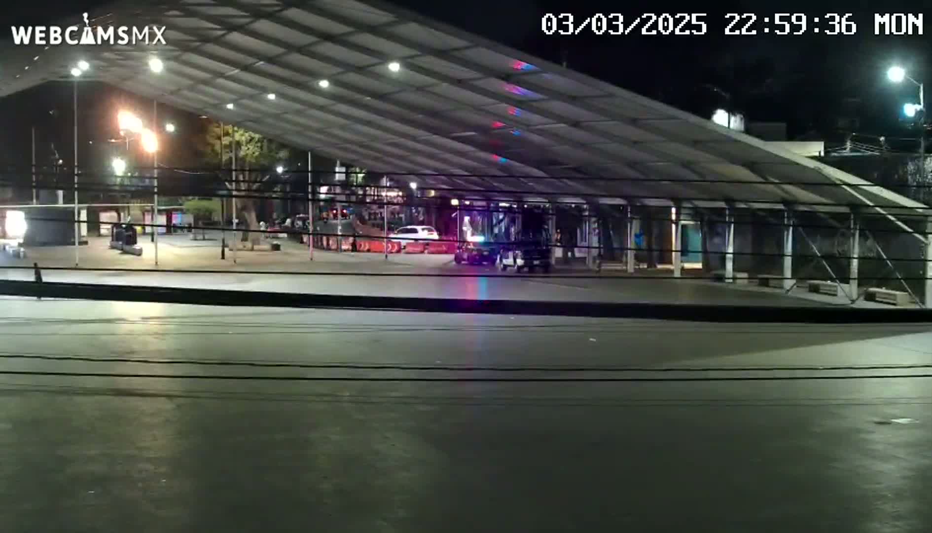 A nighttime view of a mostly empty plaza under a large covered structure, with a few cars and people visible in the distance.