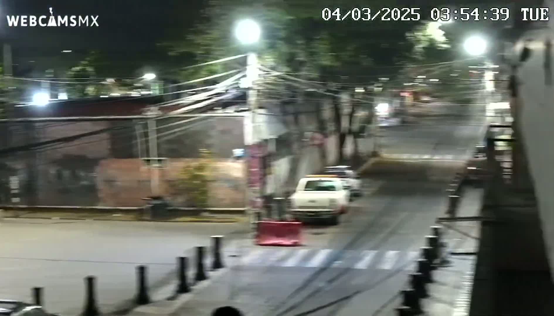 A nighttime street scene shows a mostly empty road with a white pickup truck parked near a building, under streetlights.