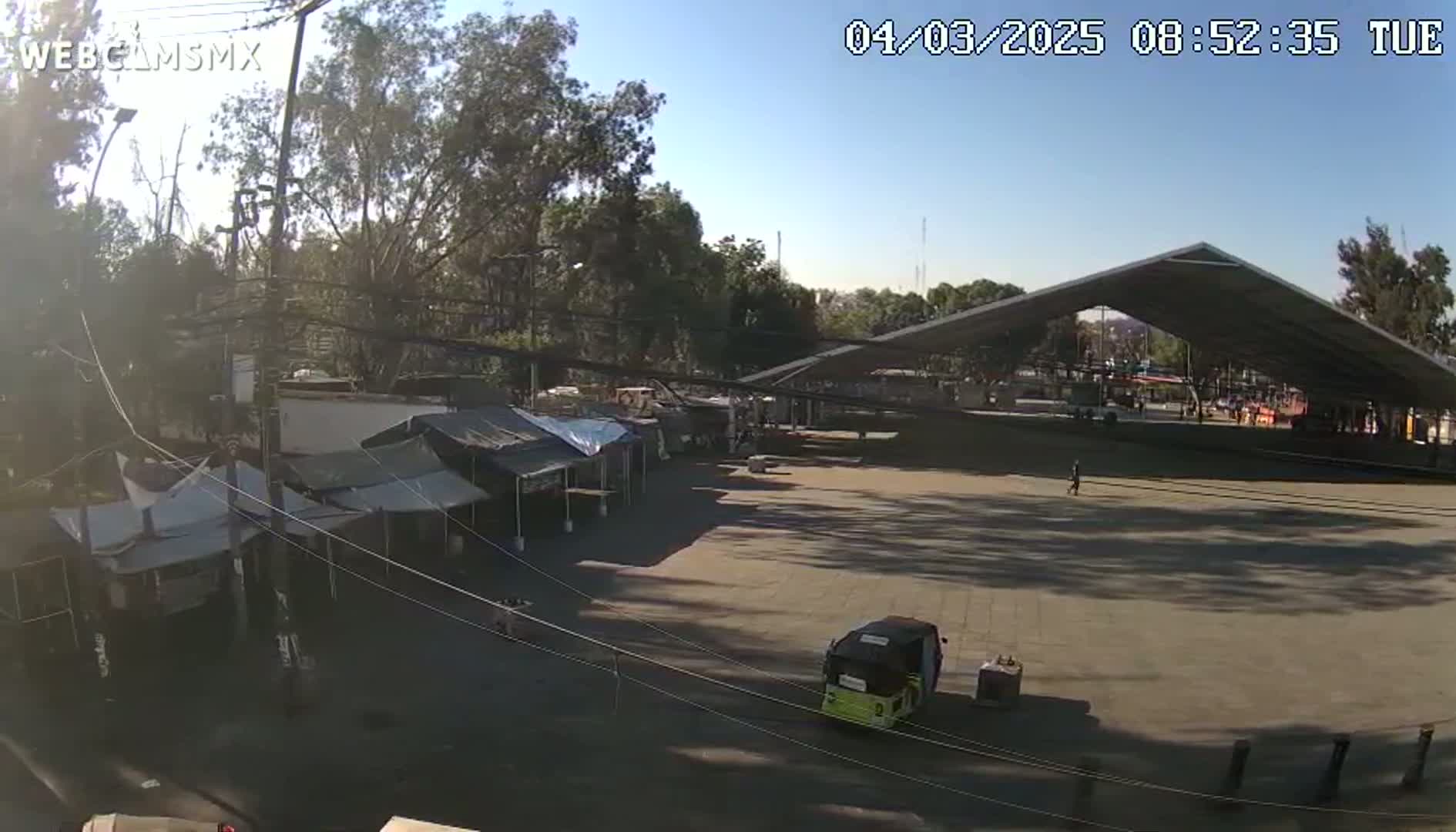 A large, empty plaza with several tents and a large covered structure is visible under a sunny sky.