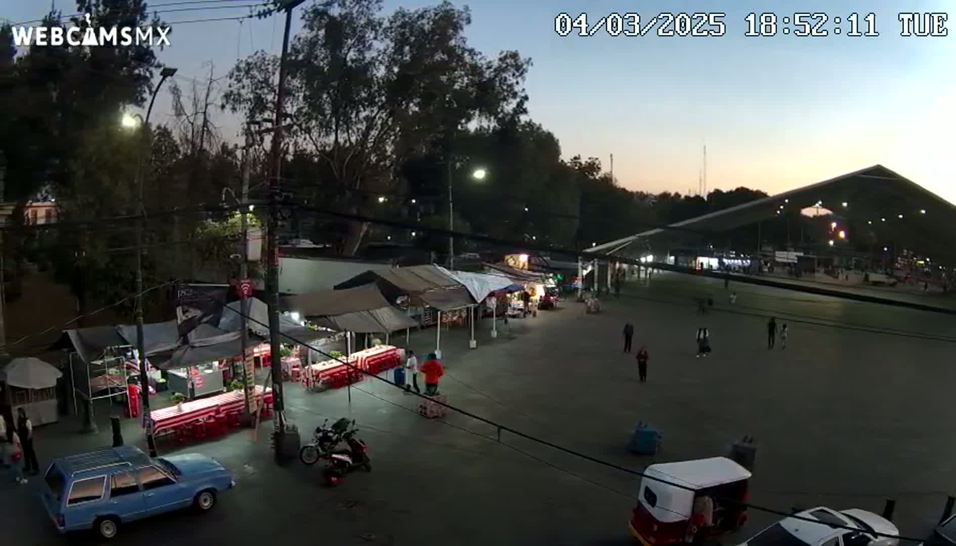 An outdoor nighttime scene shows a large paved area with several food stalls and a few people walking around, under a twilight sky.