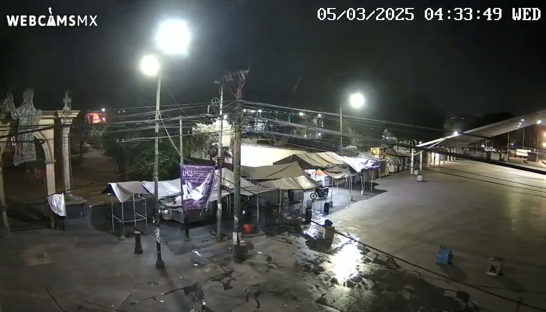 A nighttime view of a mostly empty plaza with several temporary tents, streetlights, and overhead wires, showing wet pavement.