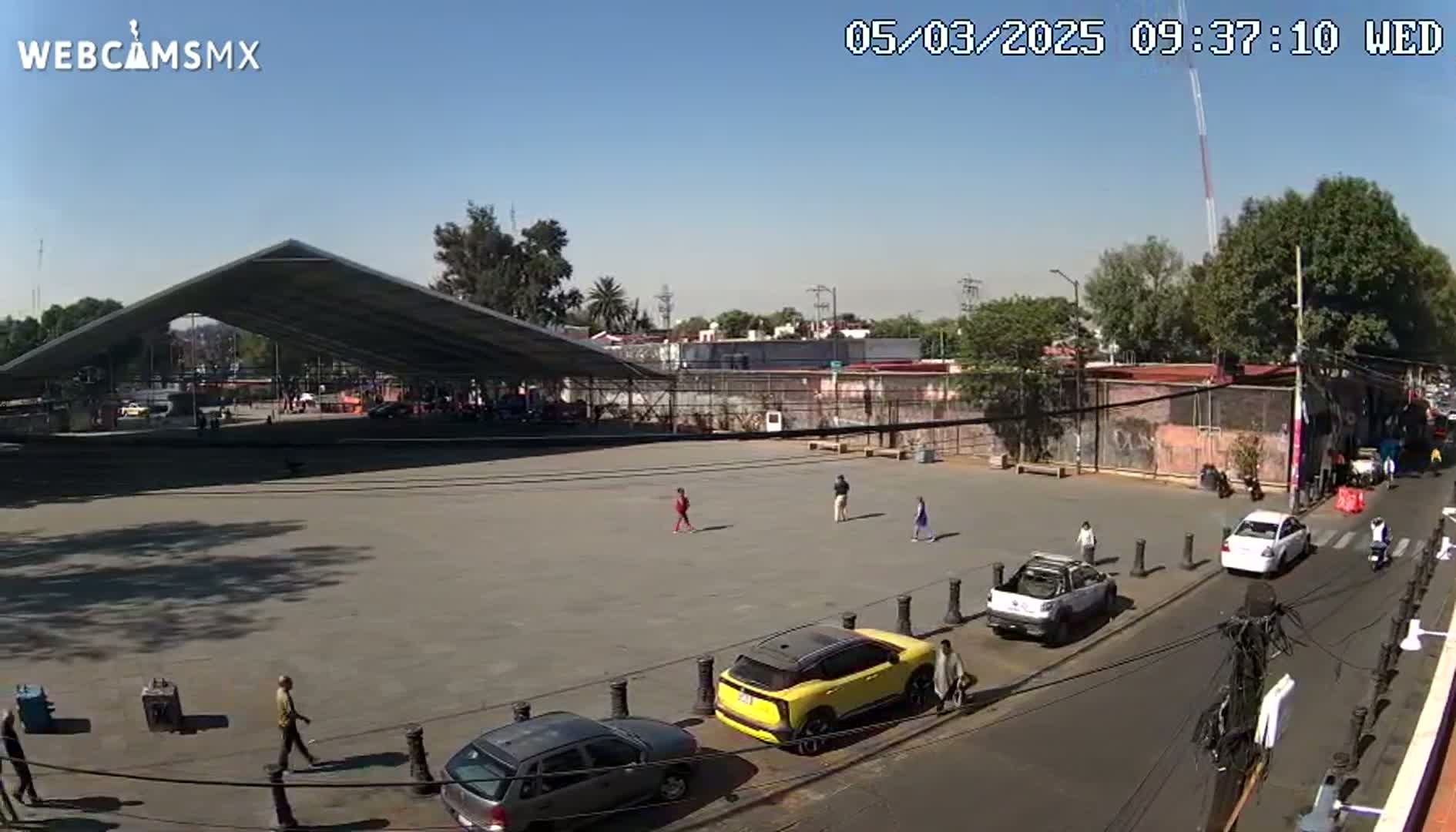 A large, open plaza with a modern, peaked roof structure sits under a clear, sunny sky, surrounded by buildings, cars, and a few pedestrians.