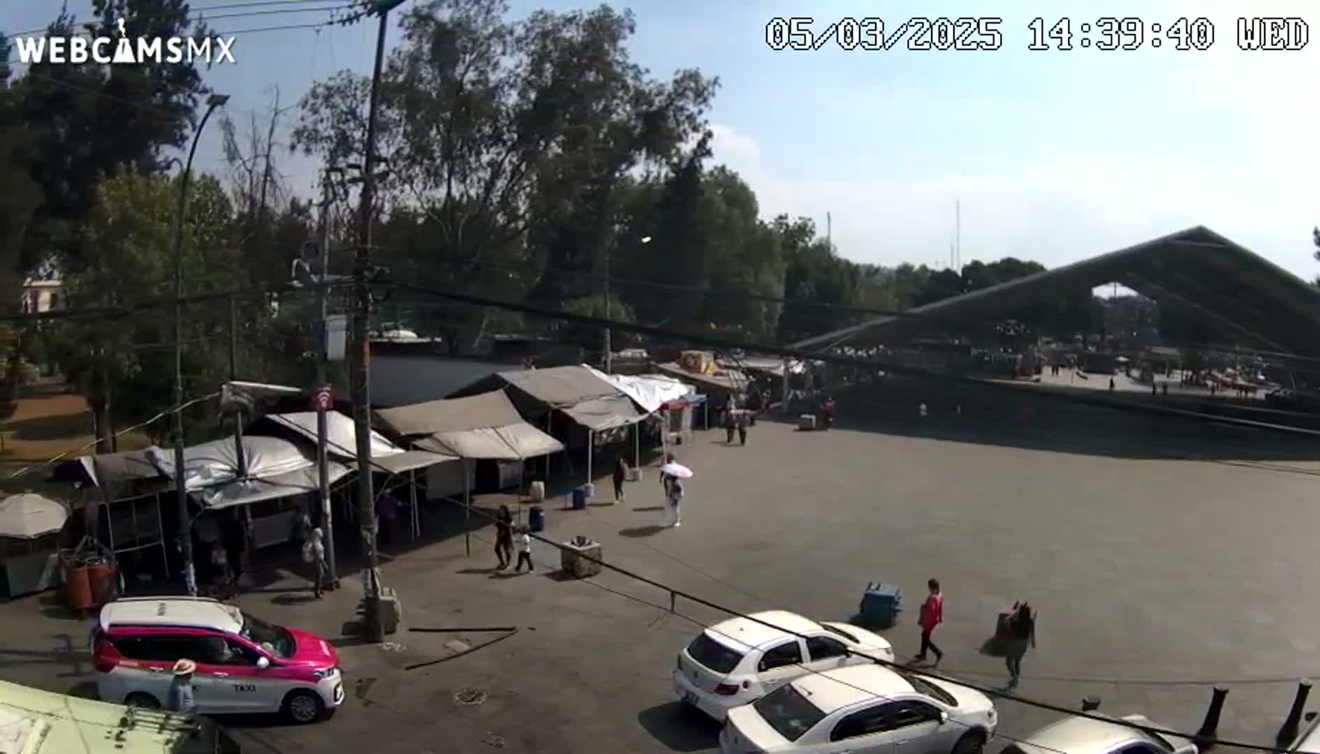 A sunny outdoor scene shows a market with numerous tents and people, along with several cars and a large structure in the background.