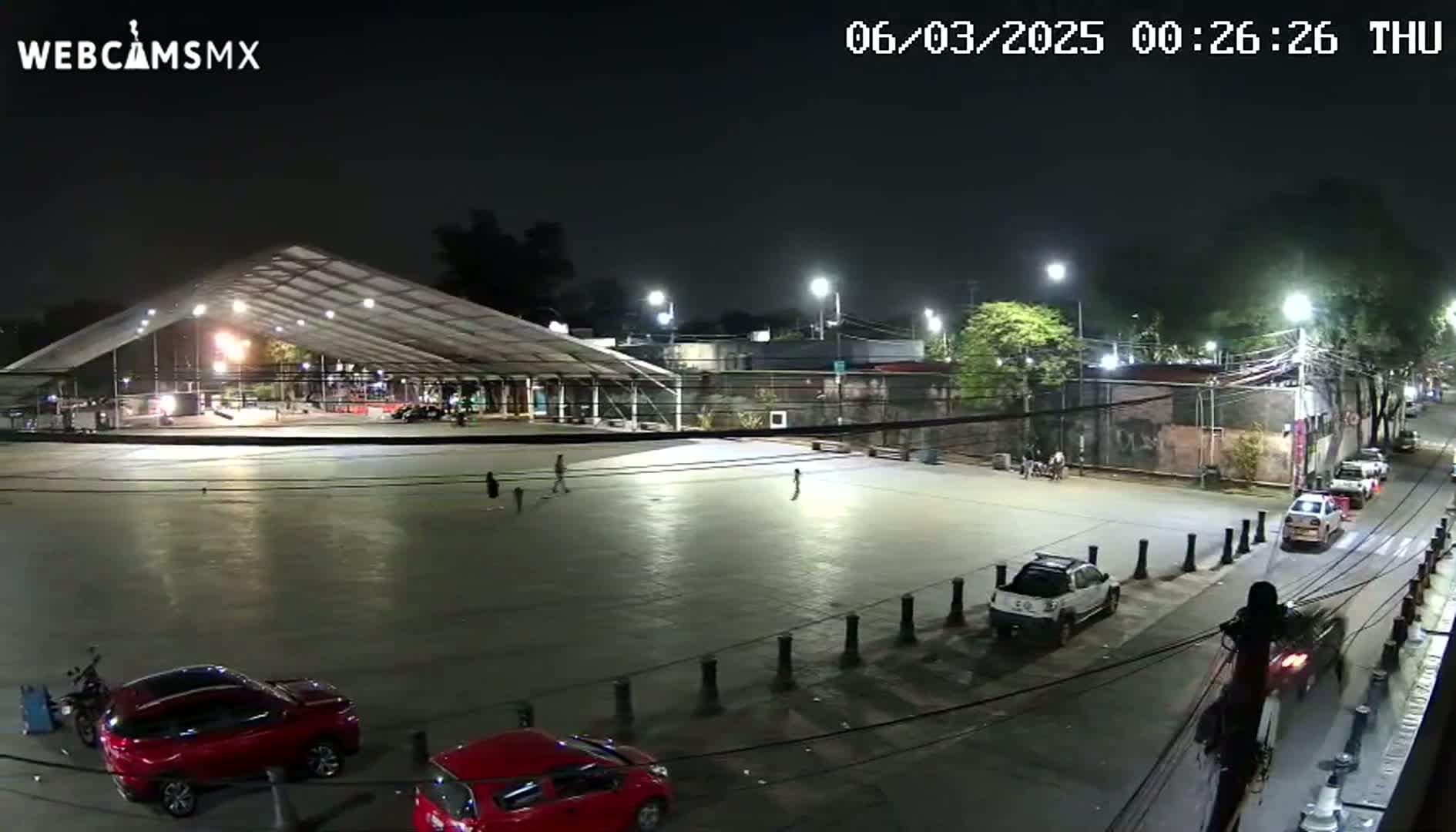 A mostly empty plaza at night, illuminated by streetlights, features a large covered structure, several parked cars, and a few pedestrians.
