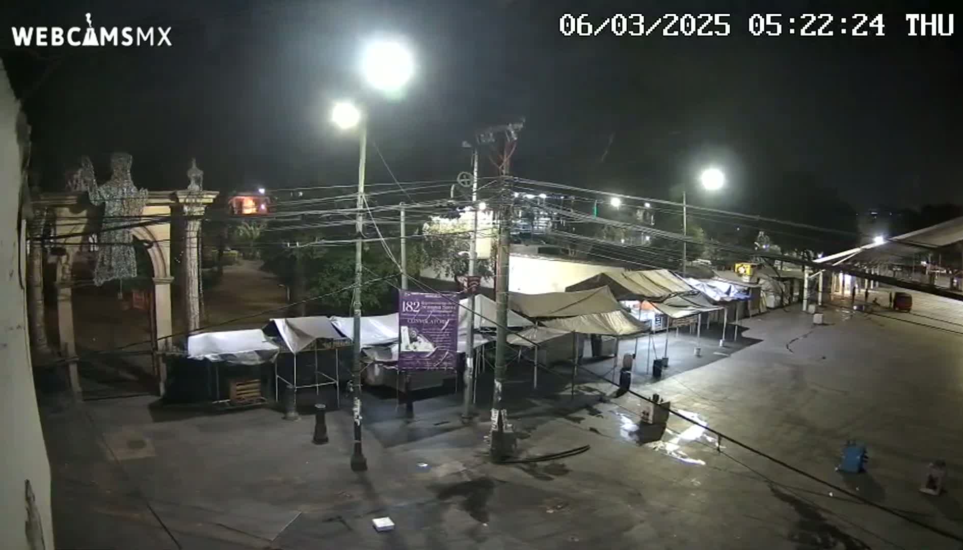 A nighttime view of a plaza with several market stalls set up under temporary canopies, illuminated by streetlights.