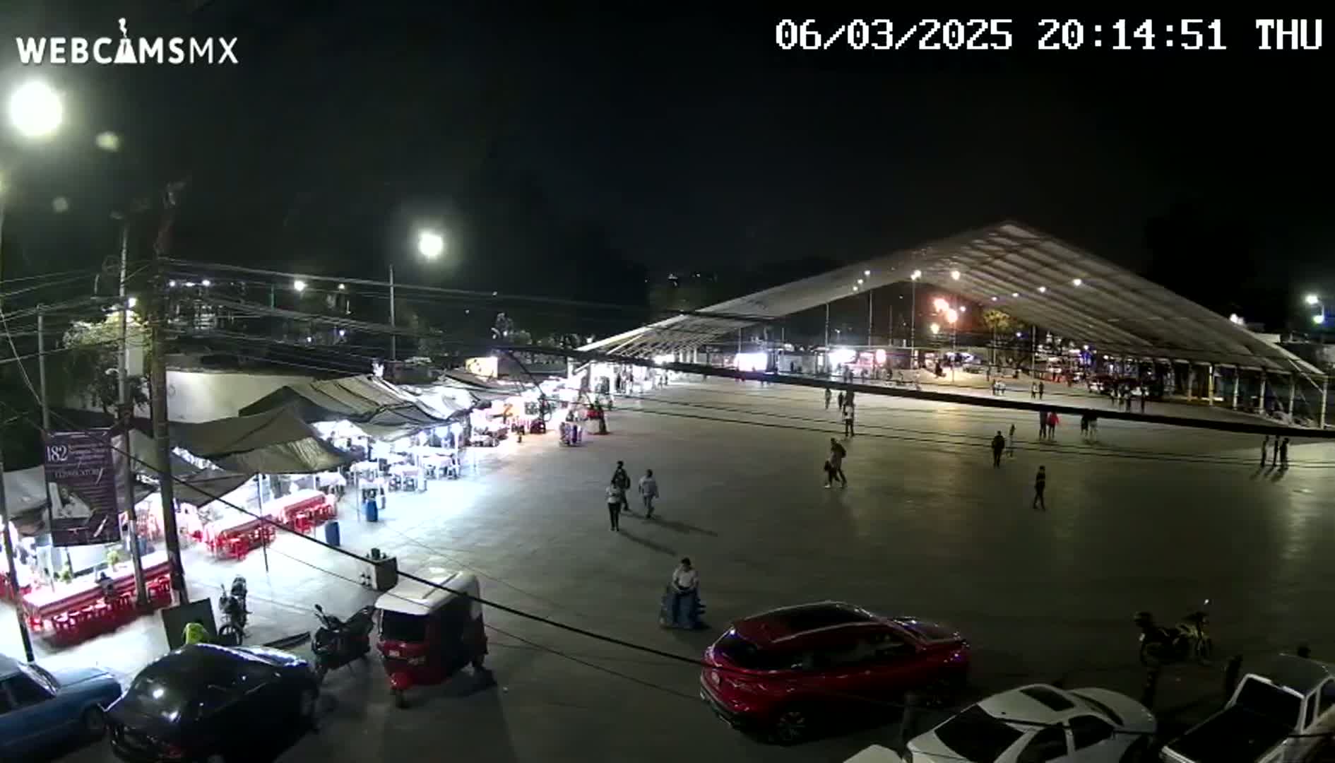 A nighttime, outdoor scene shows a large paved area with numerous food stalls and people walking, under a large white canopy structure.