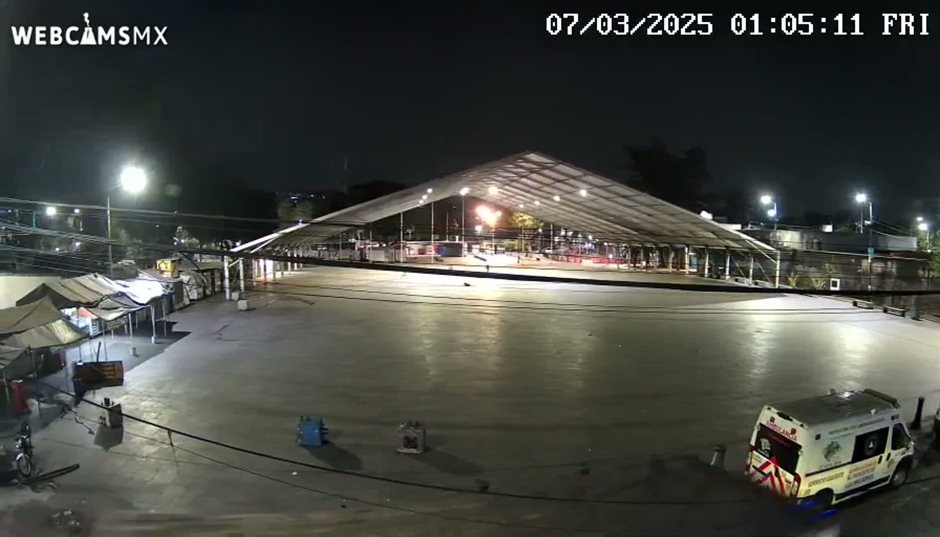 A large, empty paved area at night is illuminated by lights under a large, brightly lit tent structure, with a few small tents and an ambulance visible nearby.
