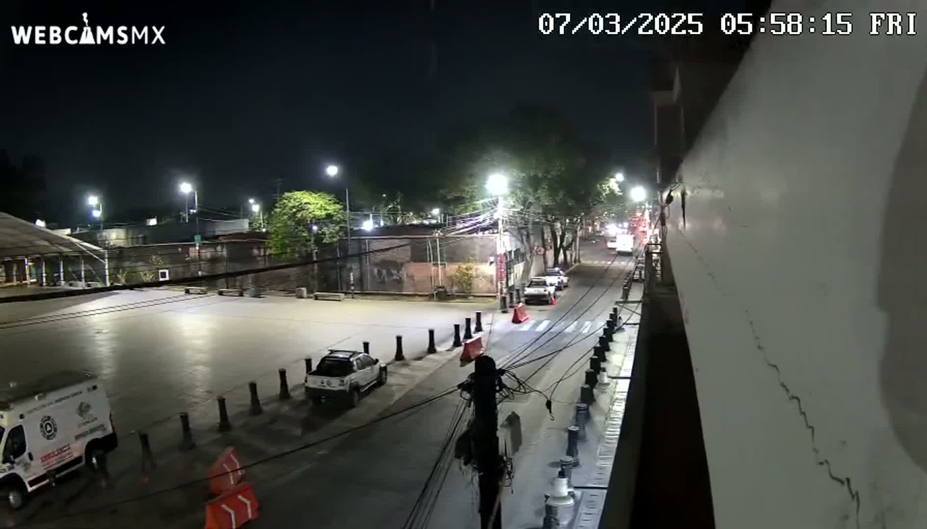 A nighttime, illuminated street scene shows several parked cars, an ambulance, and a few other vehicles traveling on a road next to a large, light-colored wall.