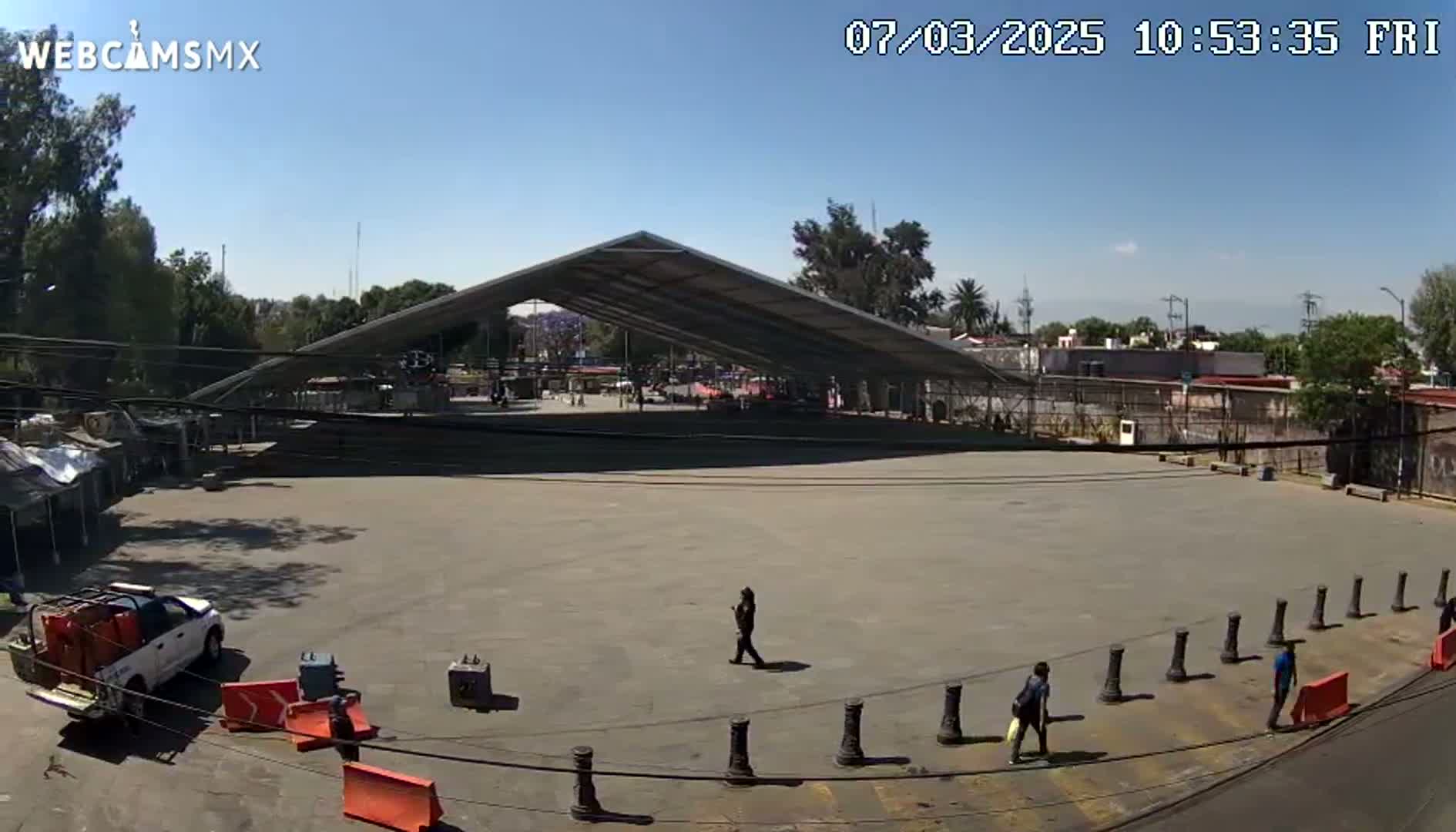 A large, empty paved area sits under a metal-framed canopy on a sunny day, with a few pedestrians and a pickup truck visible.