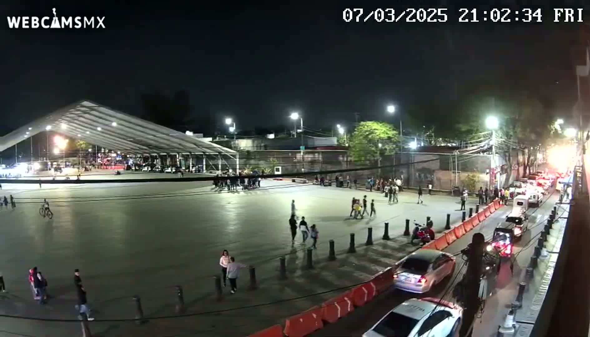 A nighttime, outdoor scene shows a paved plaza with people walking and riding bikes, a large white tent structure, and a street with cars driving past orange and white barriers.