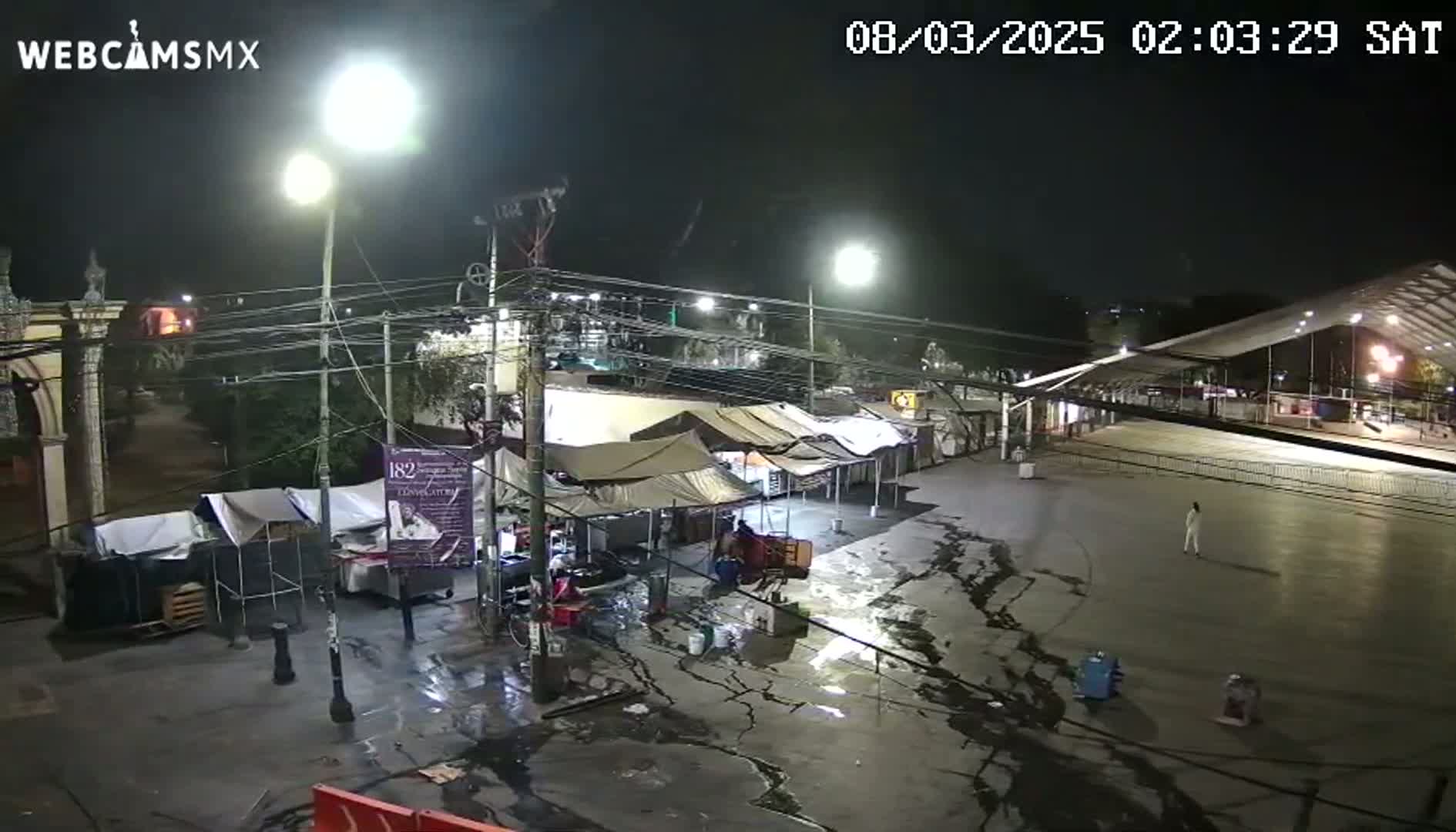 A nighttime, wet, outdoor scene shows a mostly empty paved area with several tents and a large covered structure, illuminated by streetlights.