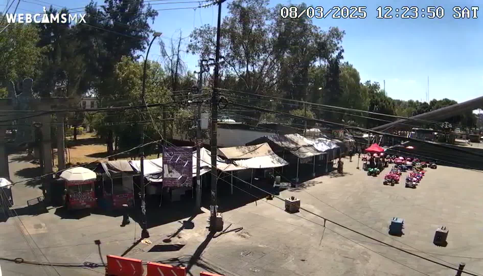 A sunny outdoor scene shows a paved area with several small tents, food stands, and a group of small cars, all surrounded by trees and power lines.