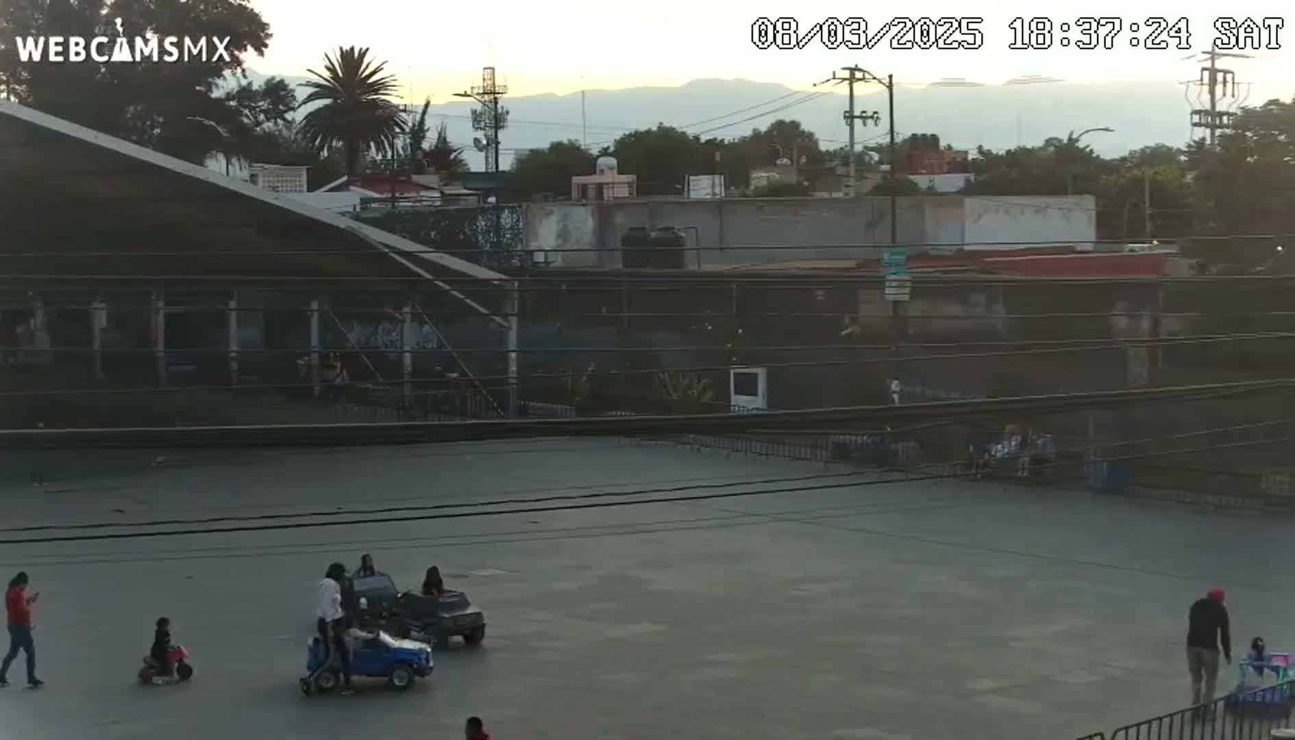 An empty paved area with several children playing in small cars under a partly cloudy evening sky.