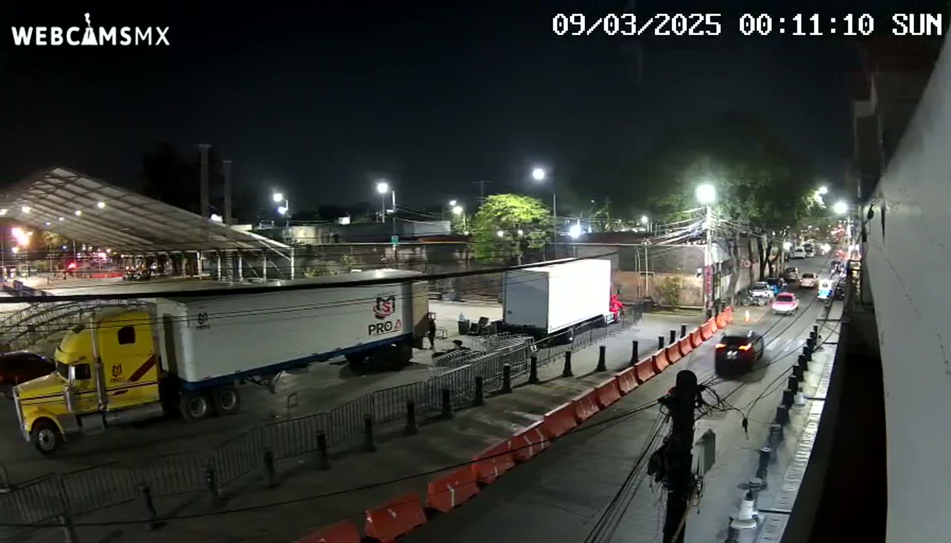 A nighttime street scene shows several trucks parked near a large tent, with a few cars driving on a nearby road under clear skies.