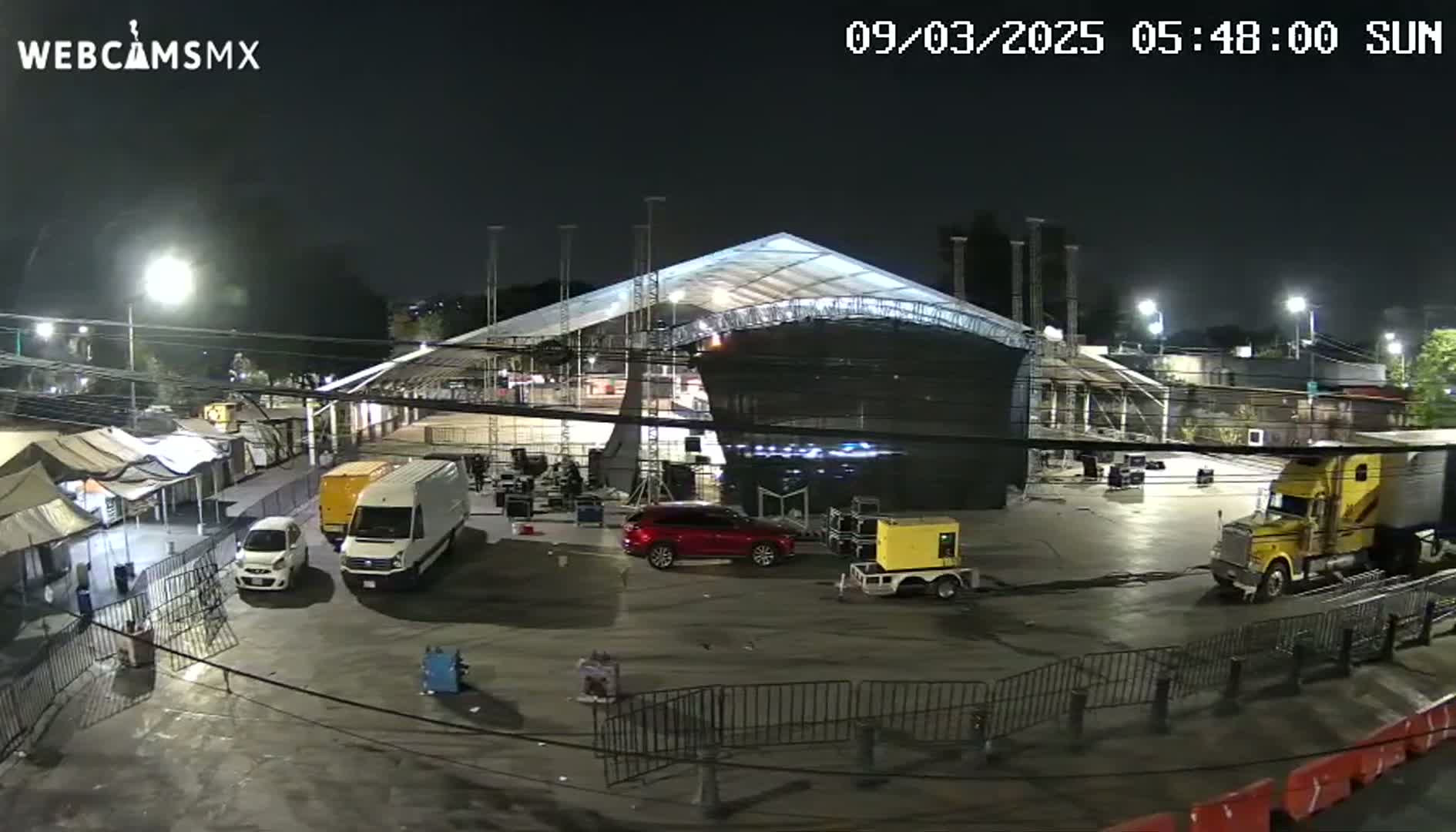 A large, temporary structure is illuminated at night in an empty parking lot with several vehicles, including a van, SUV, and semi-truck.