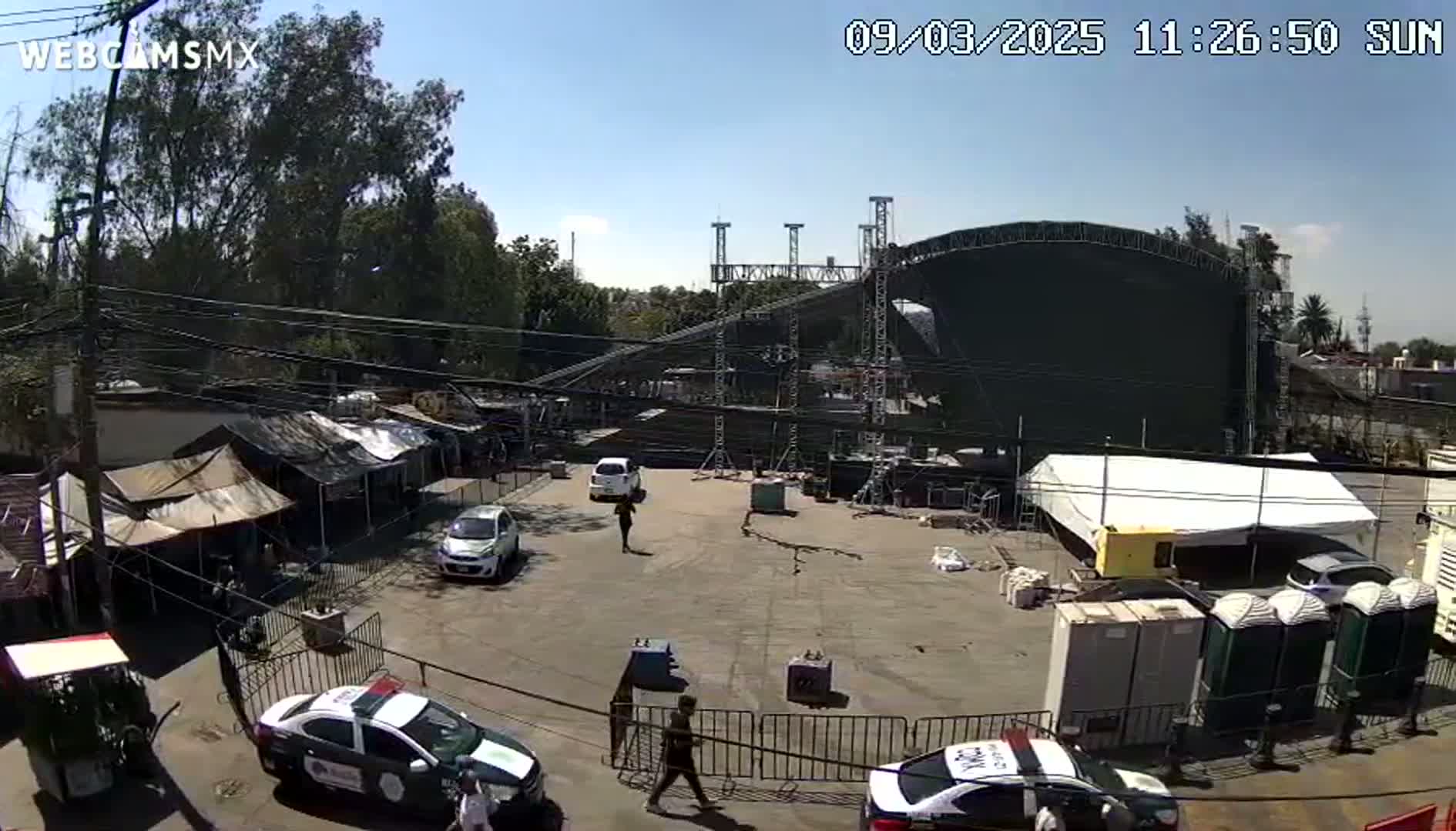 A large outdoor stage is being set up in a parking lot under a sunny sky, with several cars, people, and temporary structures present.